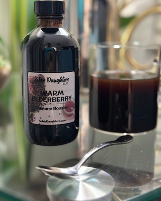 Sebi’s Daughters Warm Elderberry Tea – 8oz bottle of limited edition herbal tea blend placed beside a steaming glass of brewed tea, with a spoon resting on the counter after stirring, ingredients visible in the rich-colored tea